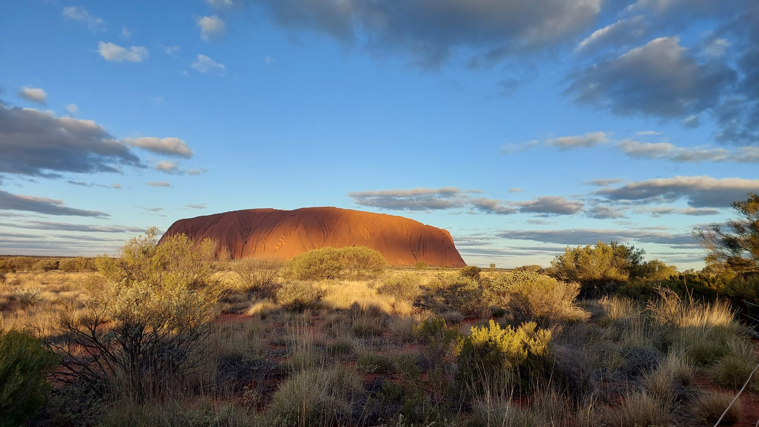 Red sandstone rock formation rises from a desert plain with sparse shrubs under a partly cloudy blue sky.