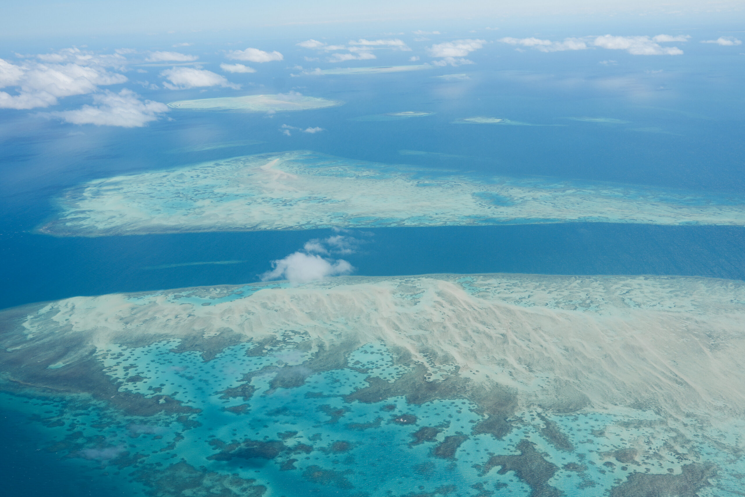 Aerial view of a turquoise tropical lagoon with scattered coral reefs and shallow sandbars near the coastline under a pale blue sky.