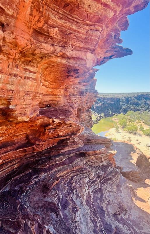 Red sandstone cliff on the left with layered rocks, a rocky ledge in the foreground, and a sunny valley with green trees and blue sky in the distance.