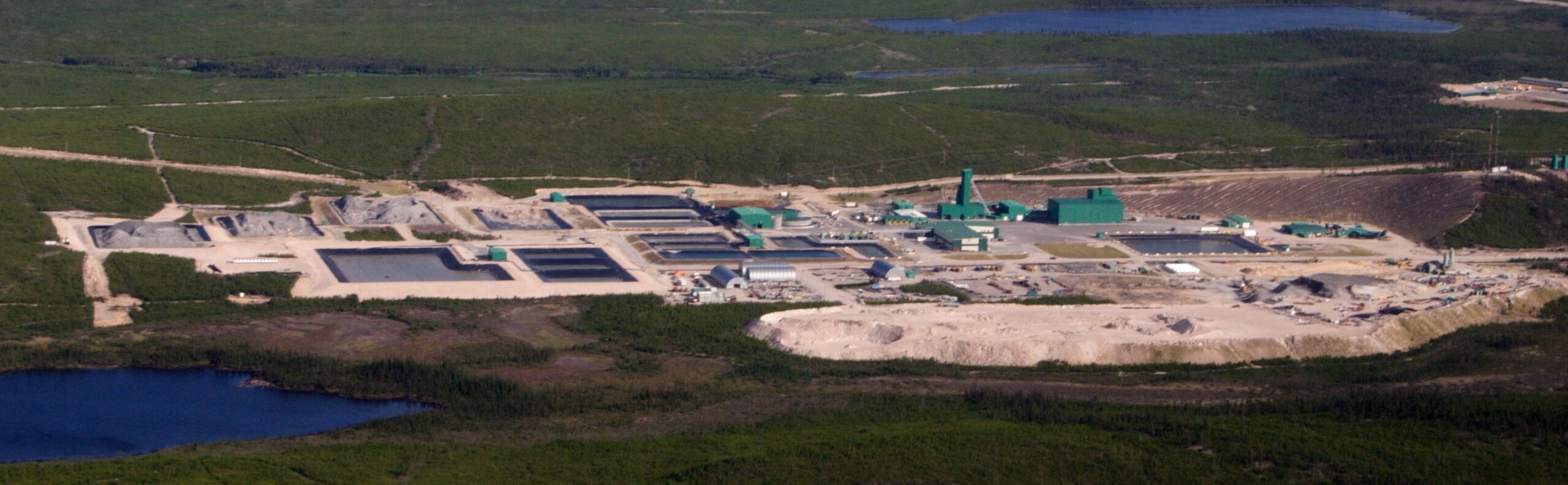 Aerial view of an industrial processing plant with green buildings and open pits, set in a green landscape near a small blue lake in the foreground.