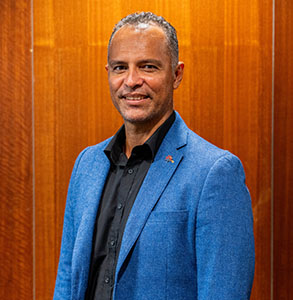 Man in a bright blue blazer and black shirt standing in front of a warm wood-paneled wall, smiling at the camera.