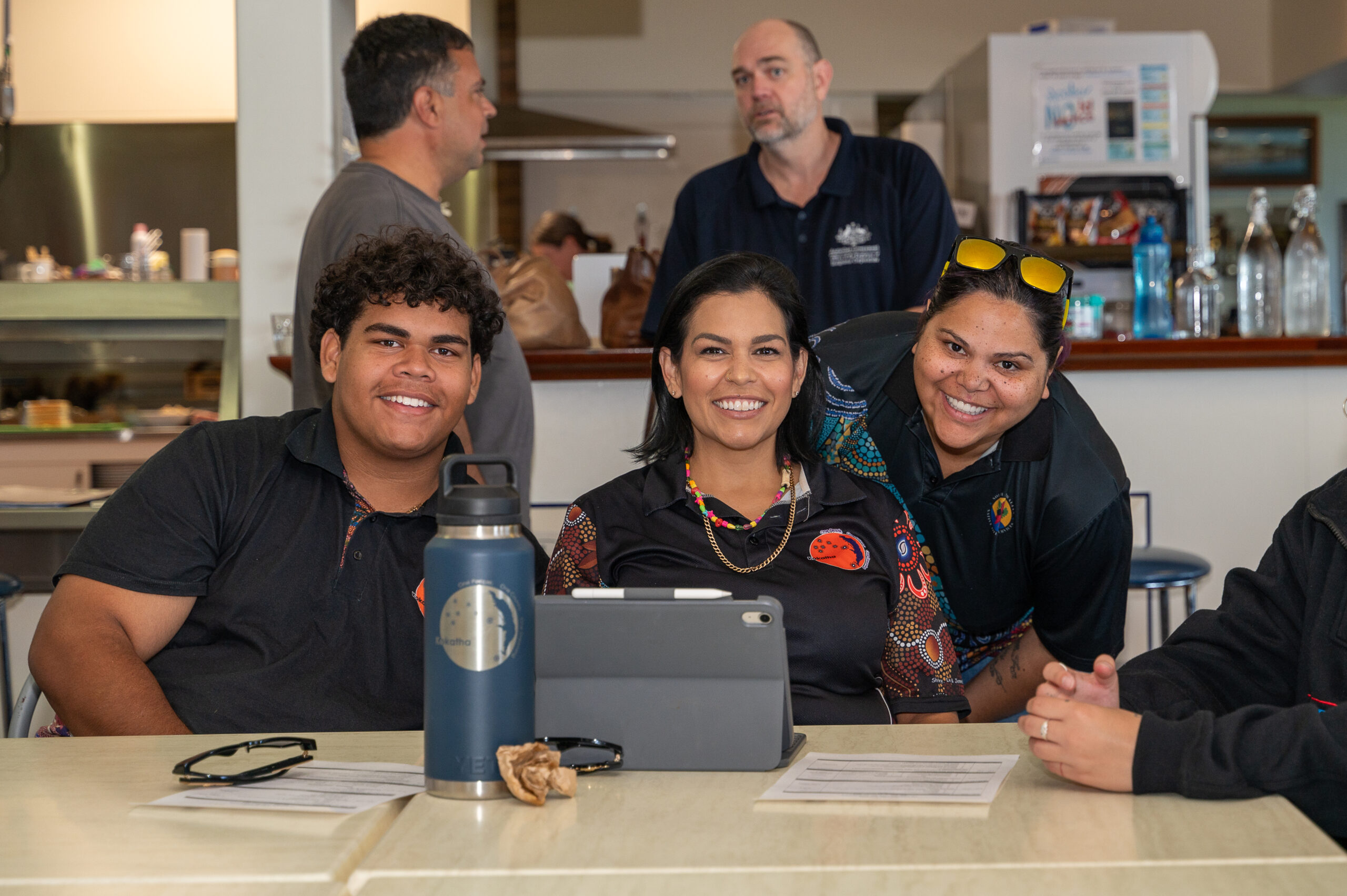 Three adults smile at the camera while seated at a cafeteria-style counter, with a tablet, water bottle, and papers in front of them.